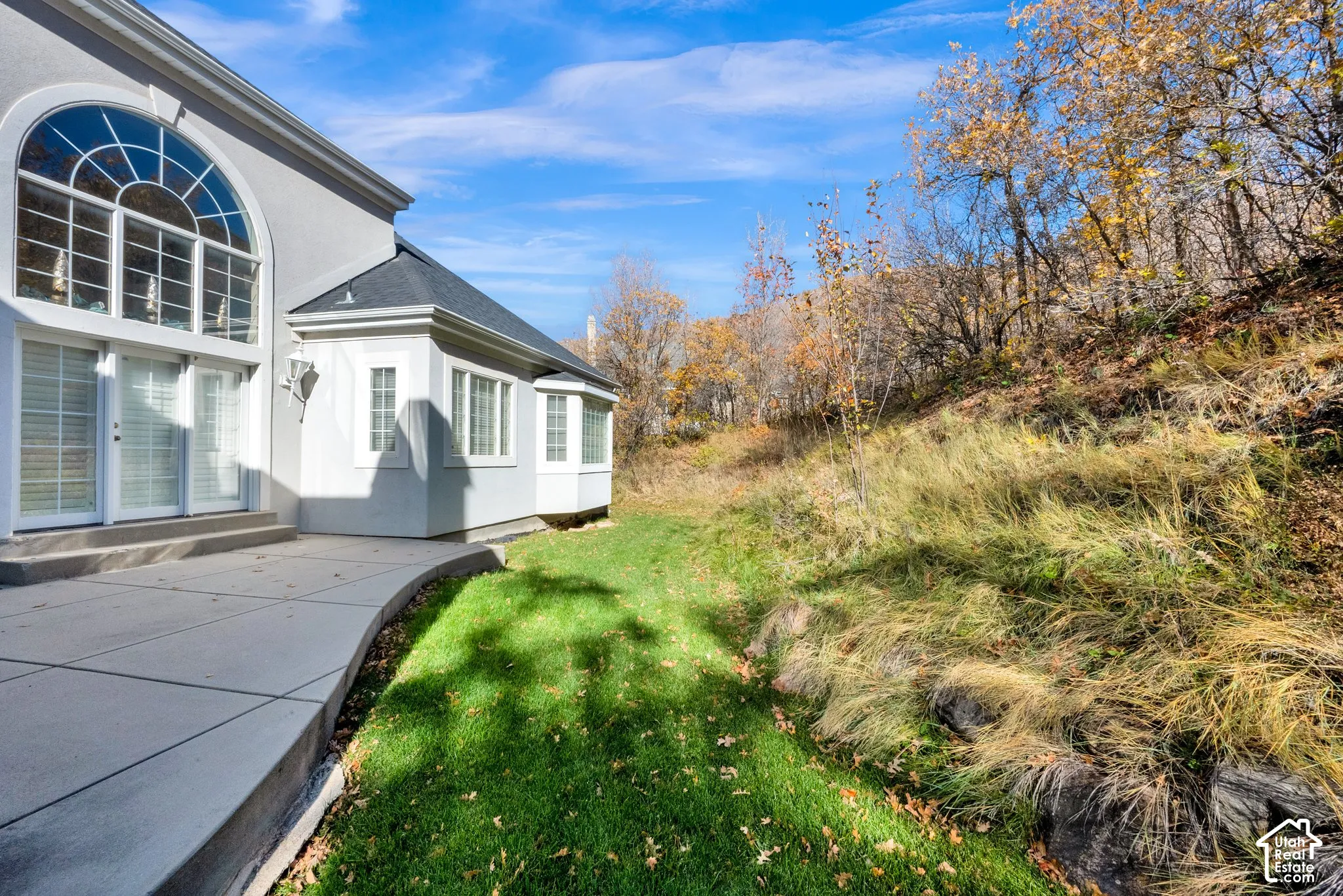 Exterior featuring a concrete patio, stucco siding, multi-pane arched windows, and a sloped rear landscape with native vegetation and natural stone terracing