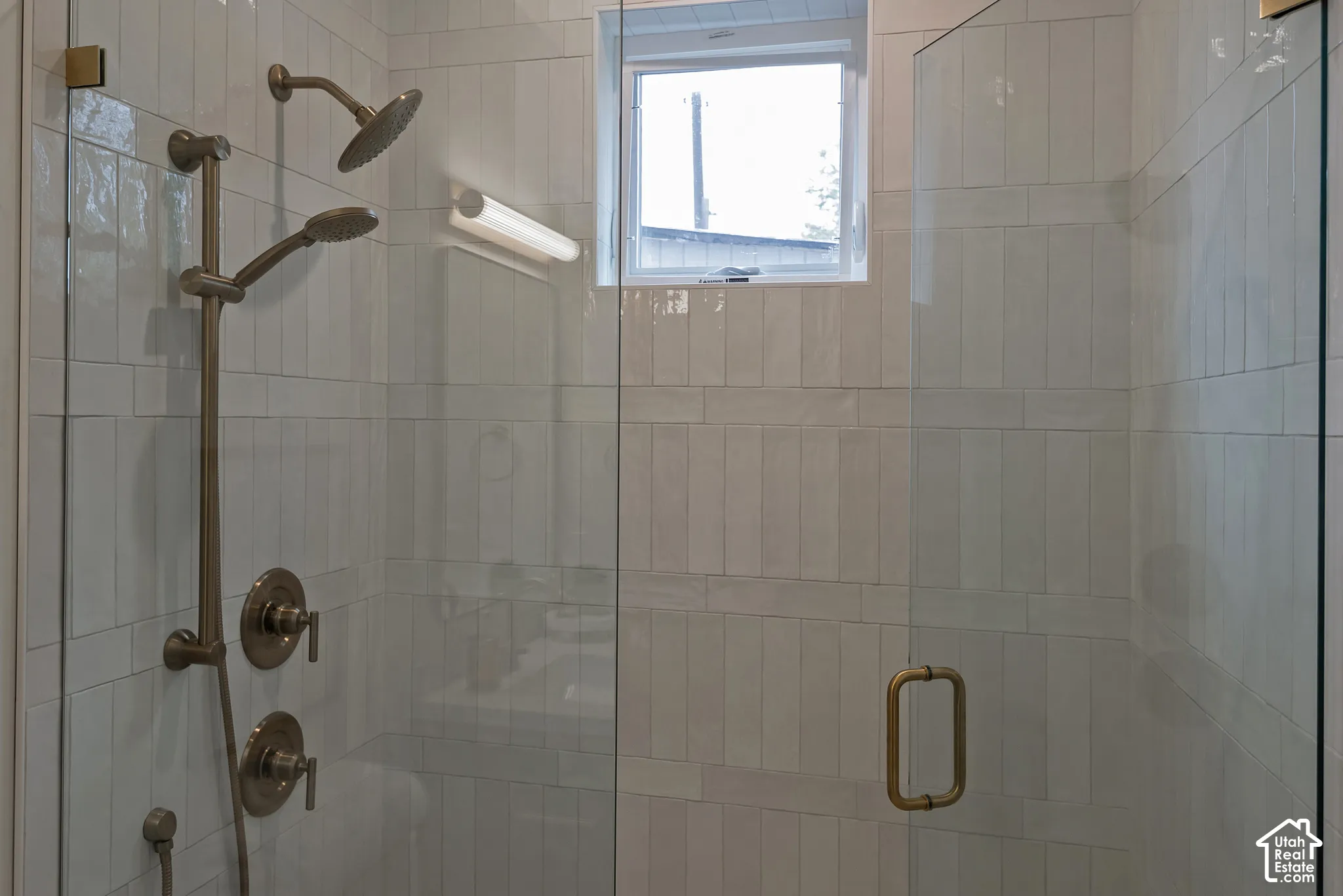 Walk-in shower featuring vertically-tiled ceramic wall surround, brass-finished dual shower heads, a frameless glass door with a matching brass handle, and a privacy window