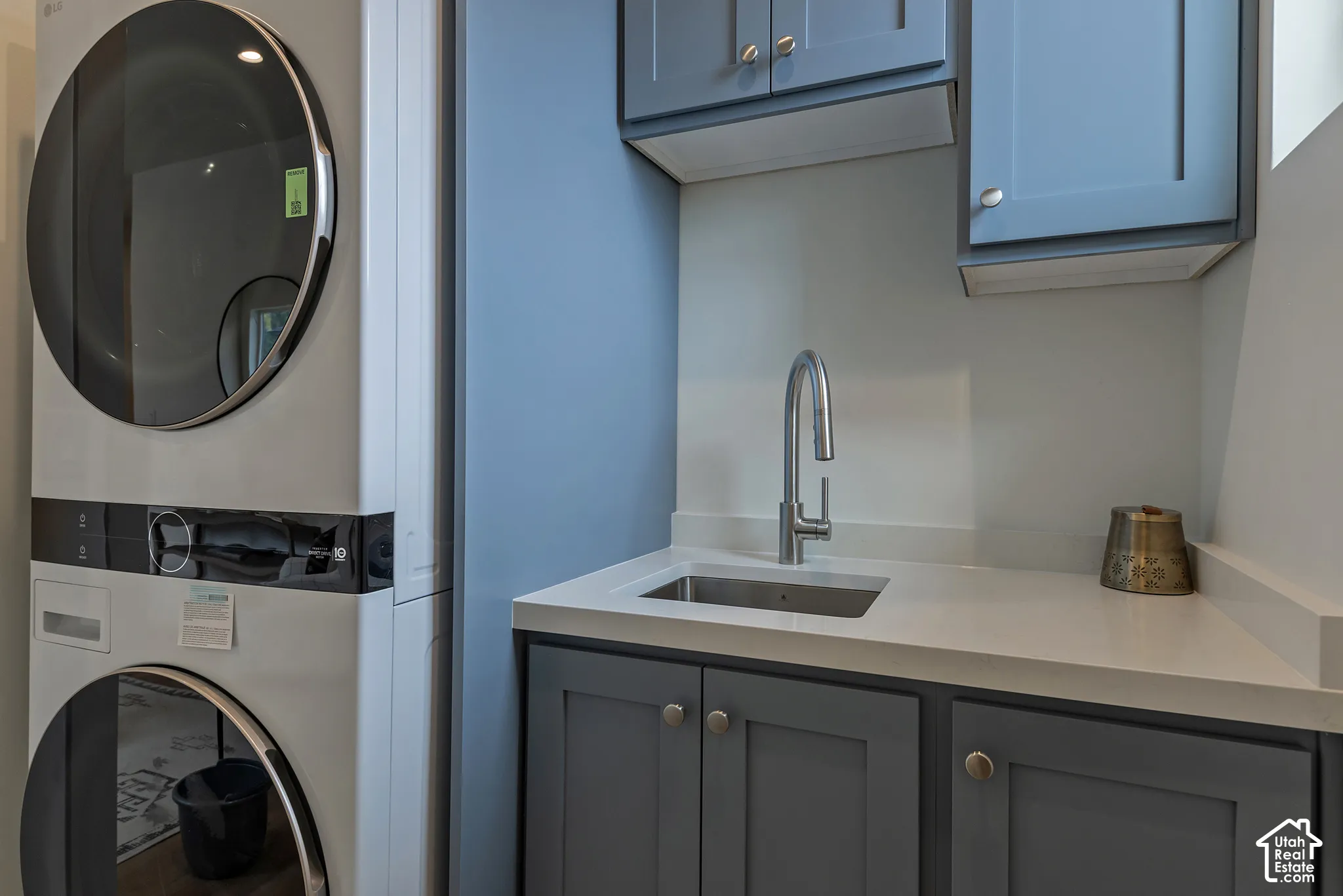 Laundry area featuring a stacked washer and dryer, solid surface countertop, undermount stainless steel sink with a high-arch pull-down faucet, and Shaker-style cabinetry