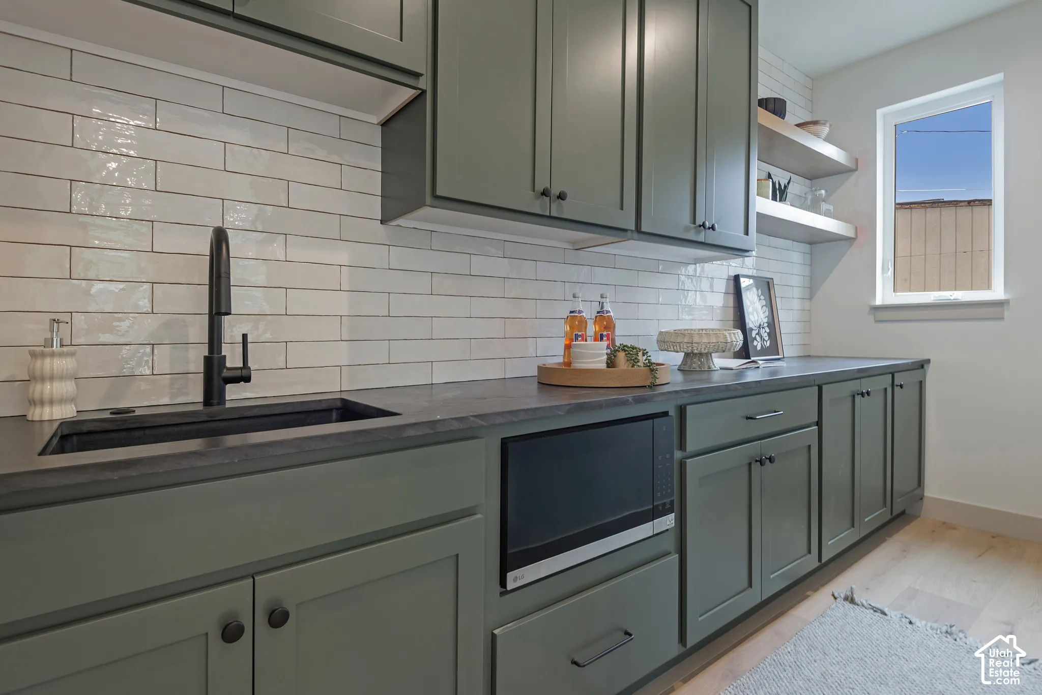 Kitchen featuring sage green shaker cabinetry, dark stone countertops, a matte black faucet with an integrated undermount sink, white subway tile backsplash, and floating wall shelves