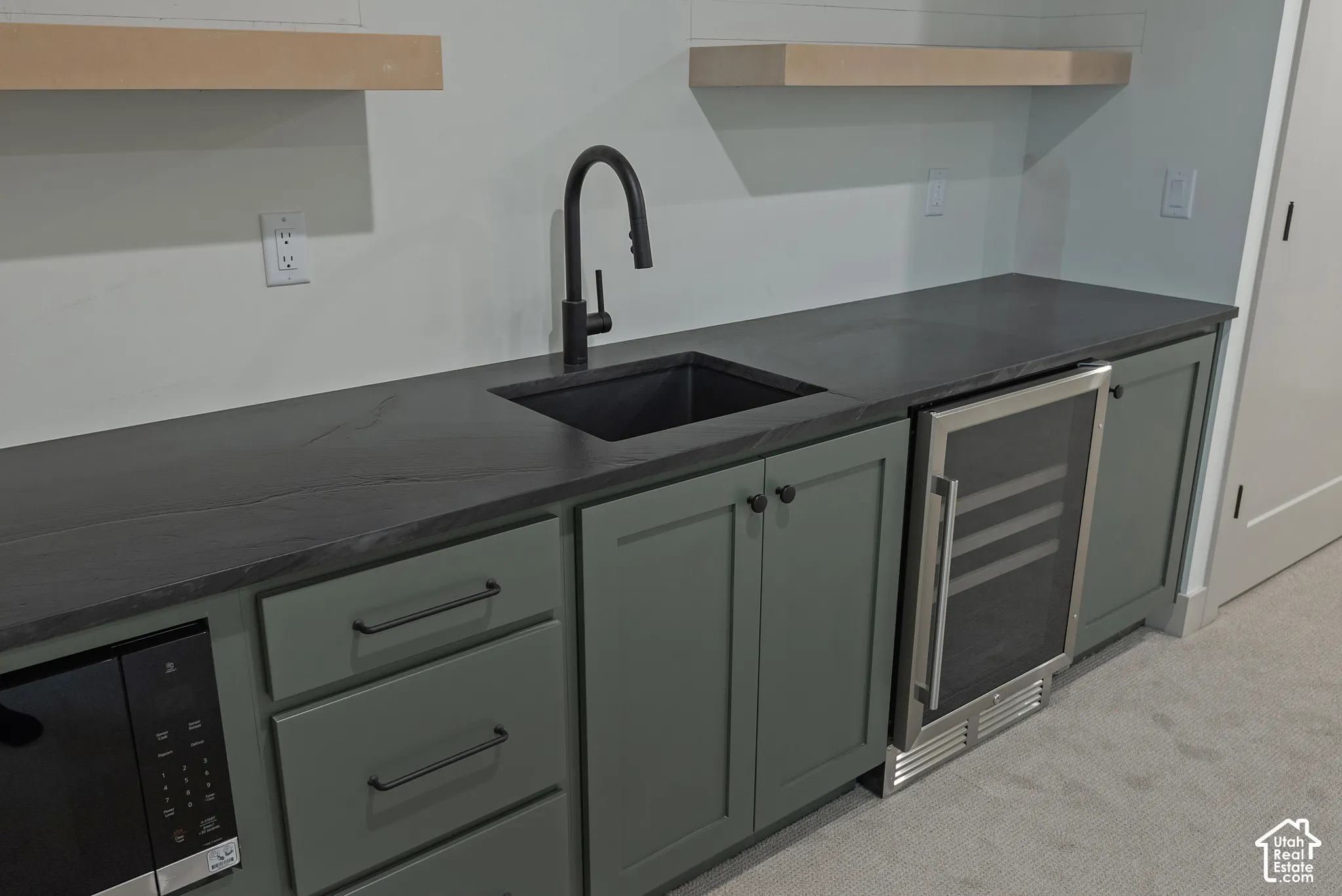 Custom cabinetry in a matte green finish featuring dark hardware, dark stone-style countertops, an undermount sink with a high-arch matte black faucet, a built-in stainless steel beverage refrigerator, and floating natural wood shelving