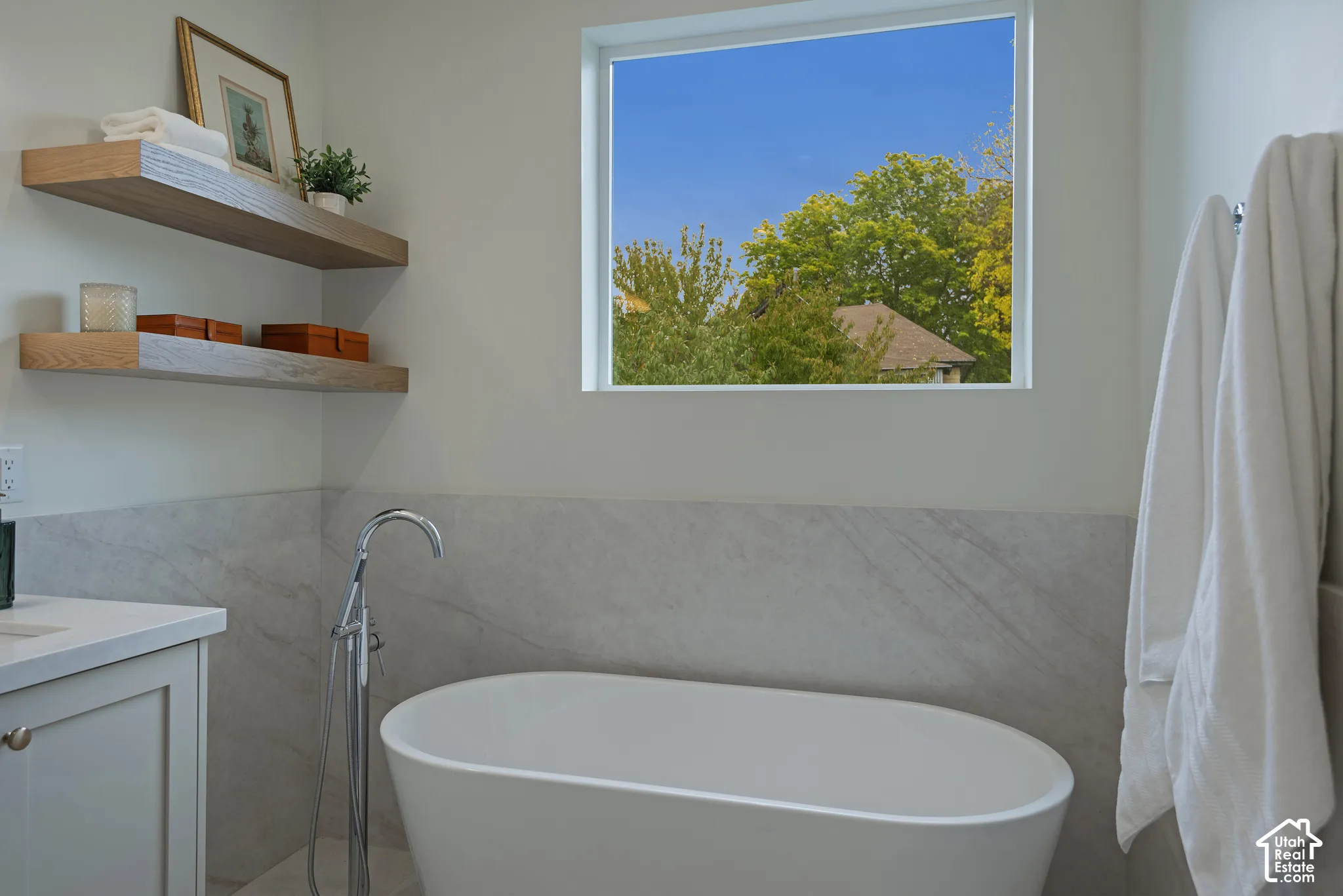 Freestanding soaking tub with a chrome floor-mounted faucet, wall-to-wall stone-finish tile surround, and dual natural wood-finish floating shelves