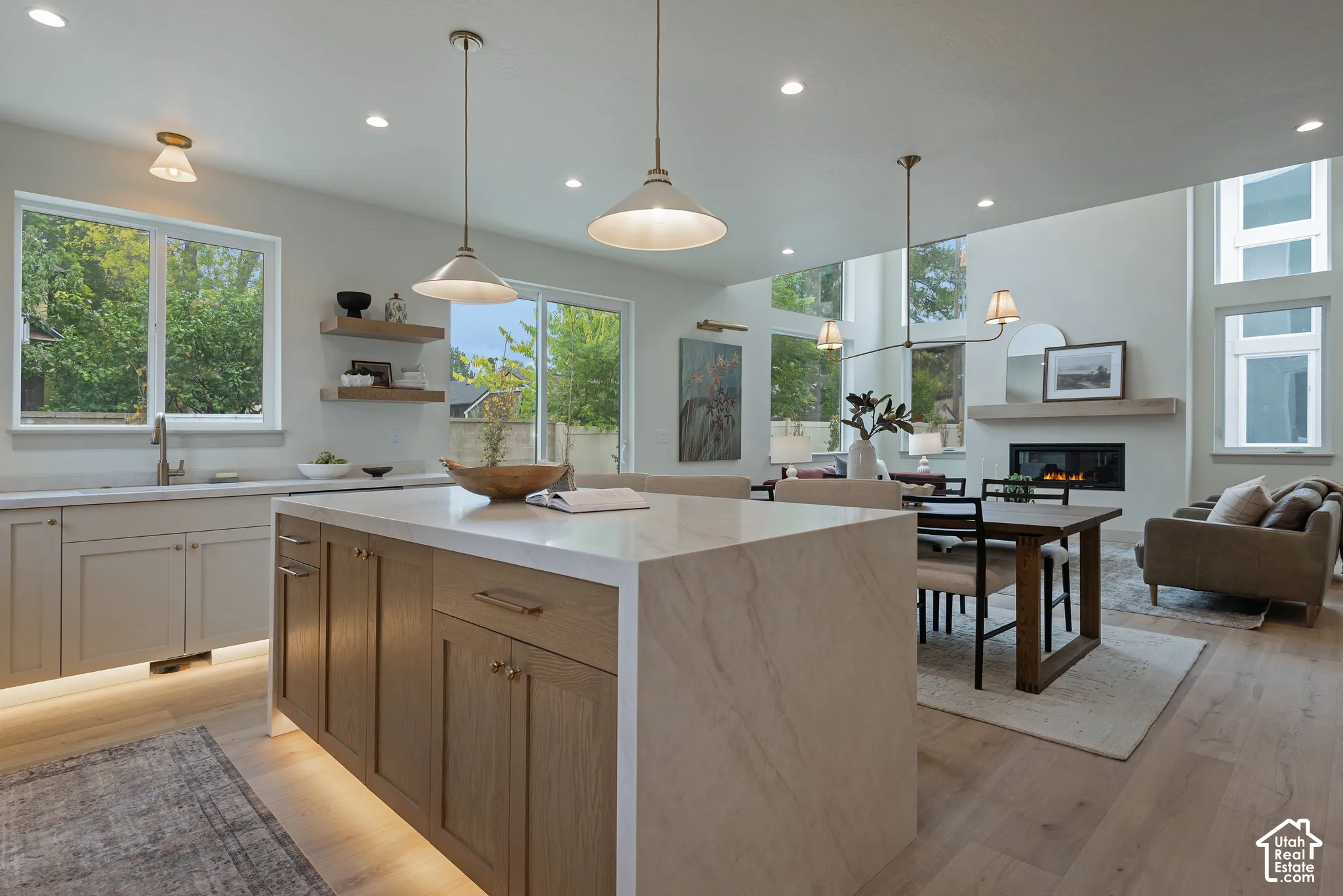 Kitchen featuring a waterfall-edge island with light-toned stone countertops, natural wood-finish cabinetry, recessed lighting, pendant fixtures, and floating wall shelves