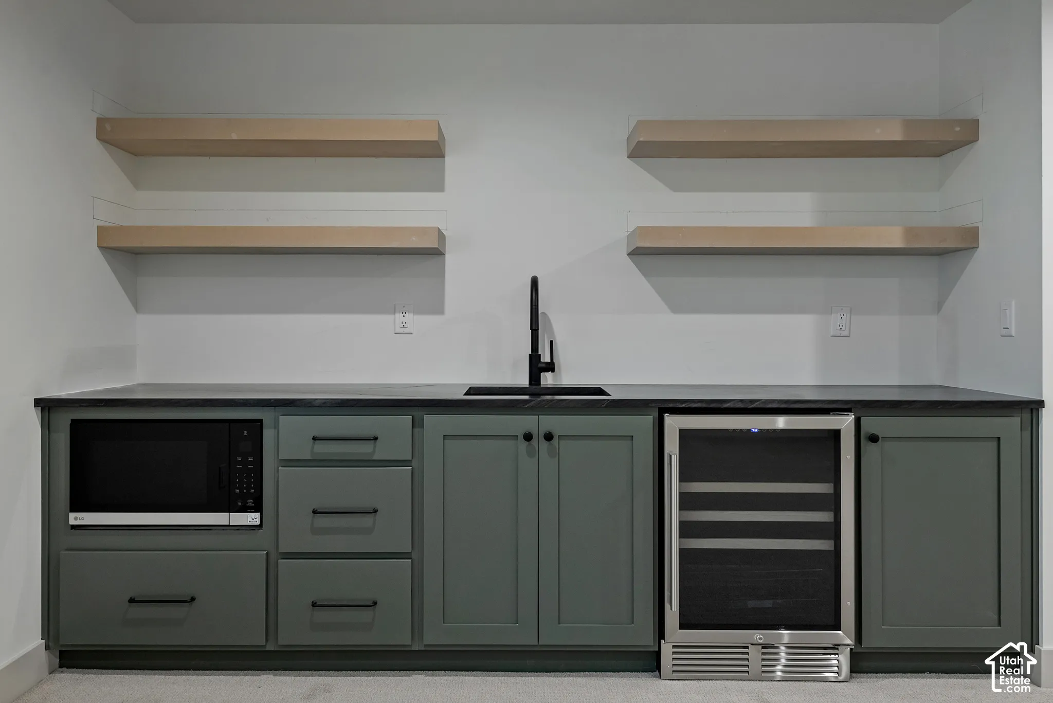 Custom wet bar featuring olive-toned shaker cabinetry, dark stone-finish countertop, integrated stainless steel beverage refrigerator, built-in microwave, and four floating wood-finish shelves with a matte black faucet