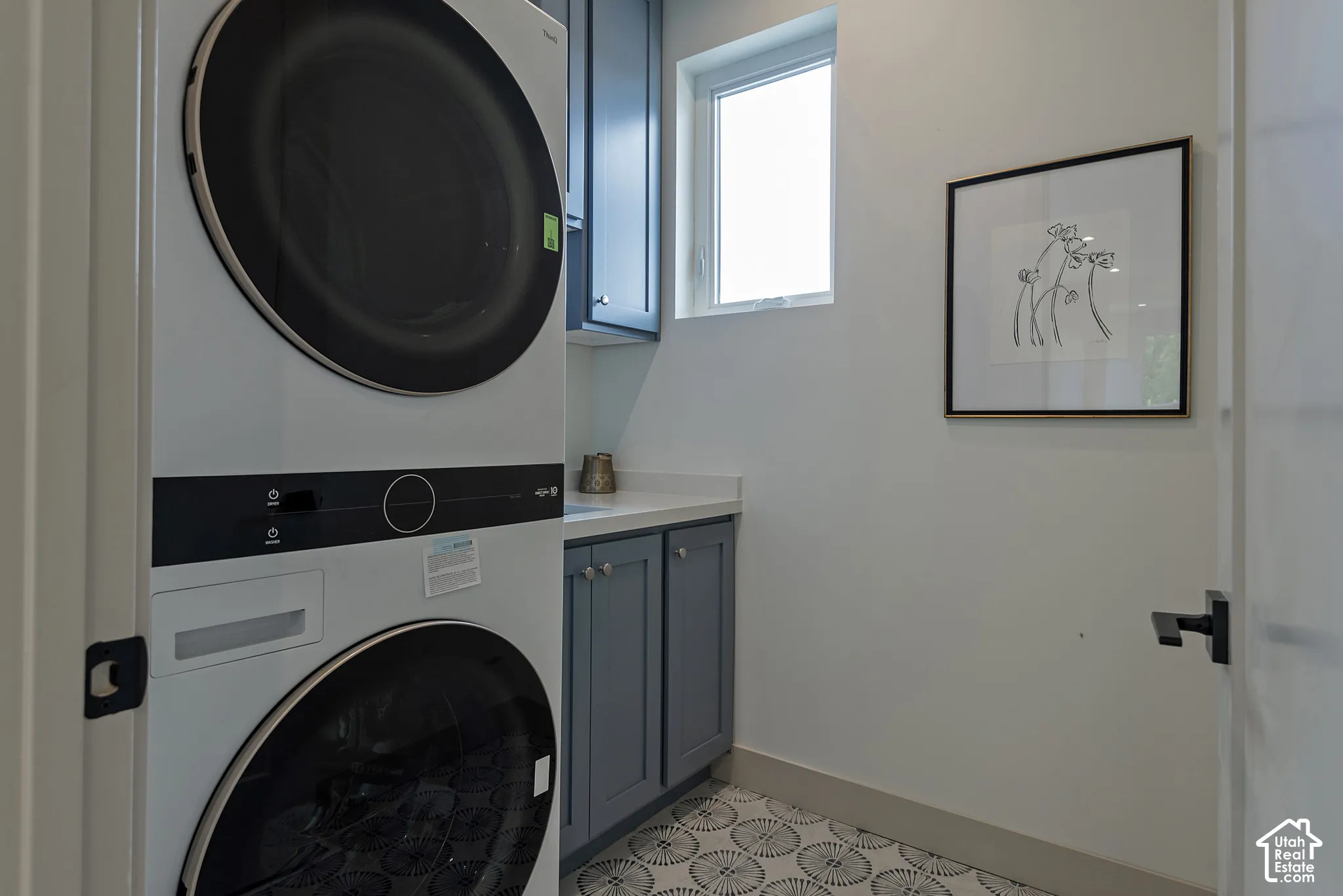 Stacked laundry appliance, blue shaker-style cabinetry, white solid-surface countertop, and patterned tile flooring