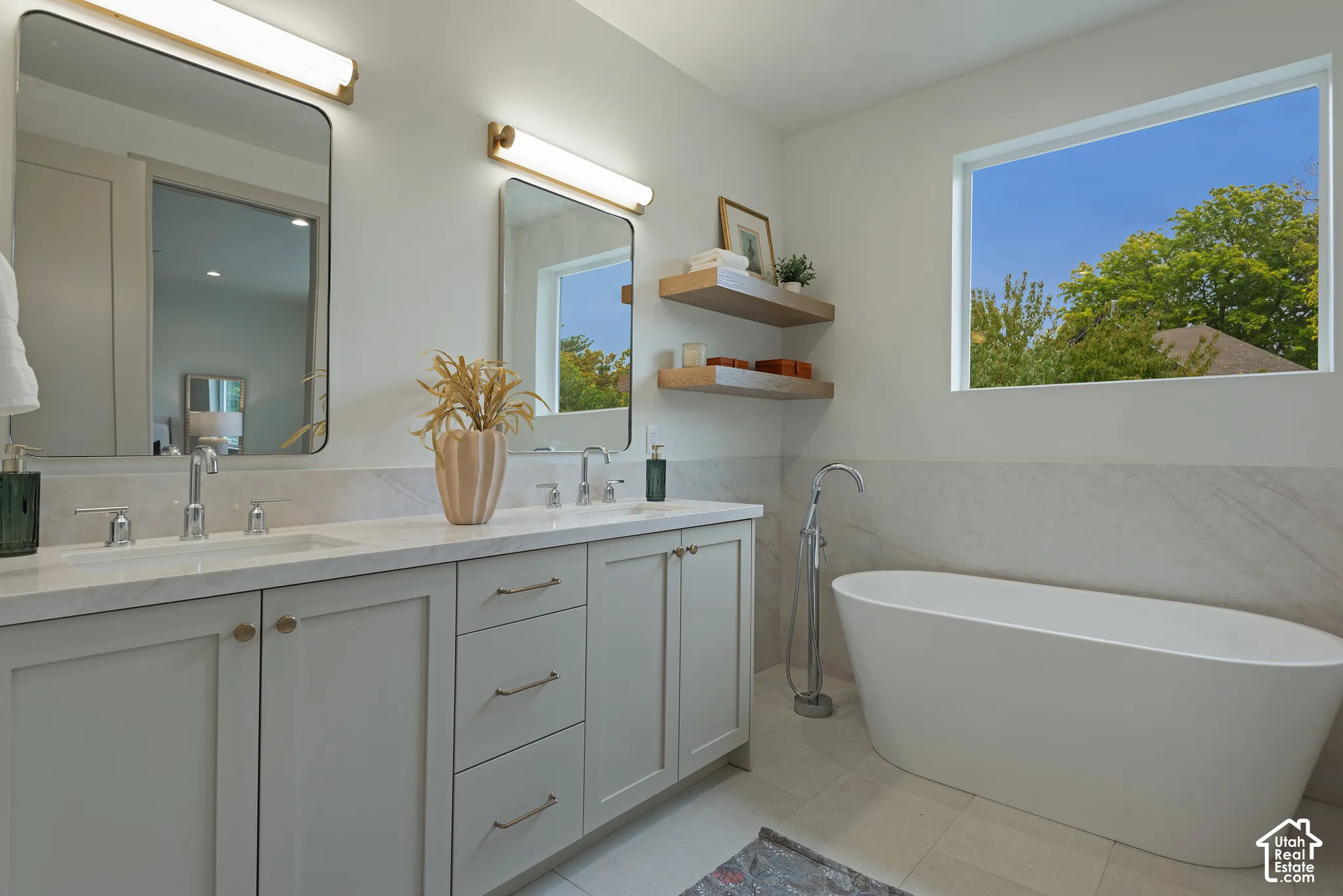 Double vanity with stone-surface countertop, freestanding soaking tub, polished metal hardware, two wall-mounted rectangular mirrors, and custom wood shelving
