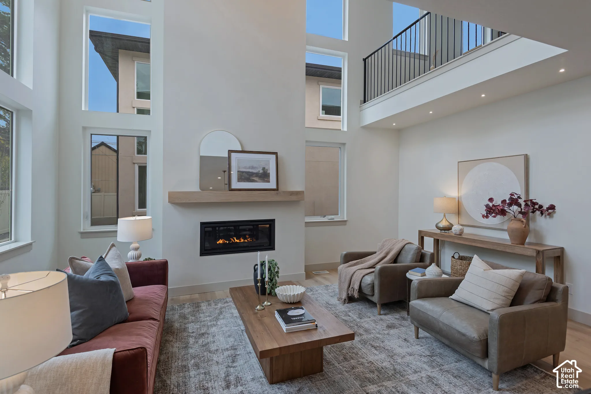 Double-height ceiling, multi-level windows, linear gas fireplace with wood mantel, and metal railing overlooking the area