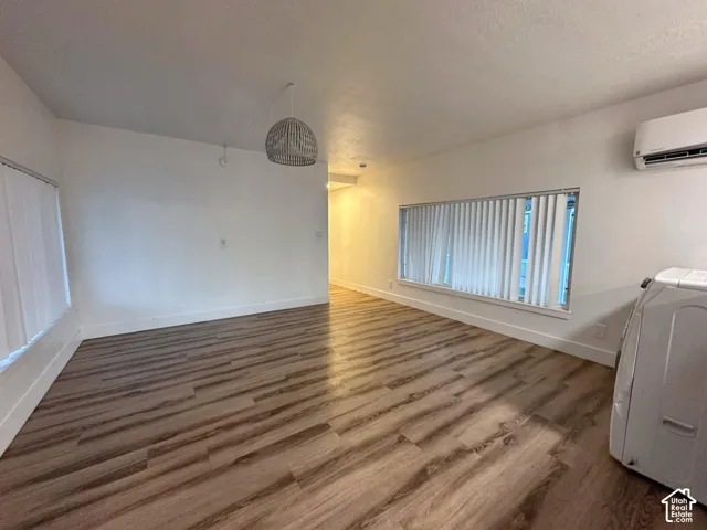 Room interior featuring wood-finish flooring, a neutral color palette, wide horizontal windows with vertical blinds, a textured dome pendant light, and a wall-mounted ductless air conditioning unit