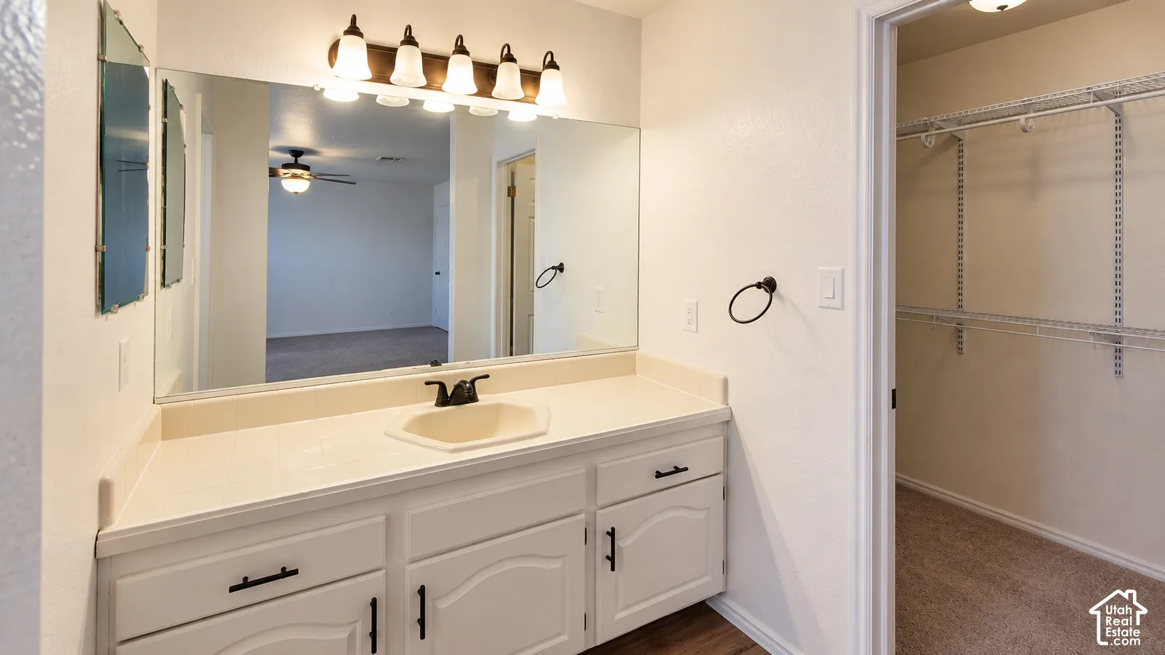 Bathroom vanity featuring white cabinetry, a neutral-toned countertop with integrated basin, matte black hardware, a multi-light fixture, and an attached walk-in closet with wire shelving