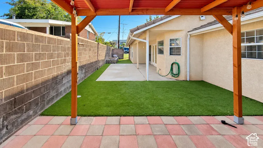 Rear exterior featuring a covered patio with a wooden pergola, concrete hardscaping, synthetic turf lawn, and a masonry perimeter wall