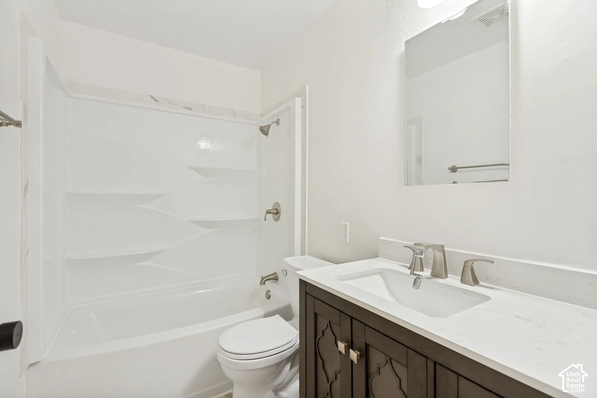 Bathroom featuring a vanity with a white composite countertop, an integrated sink, and dark-toned cabinetry with decorative paneling, complemented by a white tub-shower enclosure with built-in shelving and a frameless wall-mounted mirror