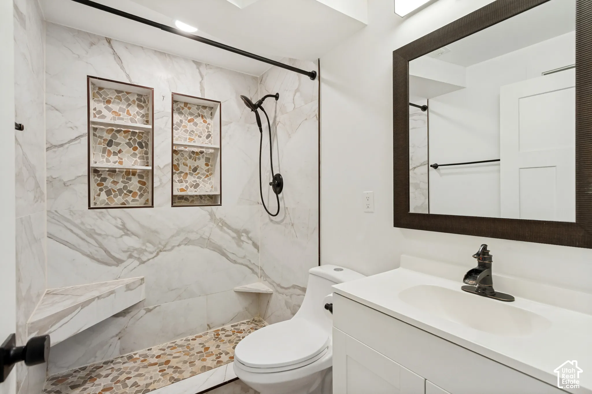 Bathroom featuring a walk-in shower with large-format tile surround, dual built-in recessed wall niches, a pebble-stone floor, and a modern vanity with a wide-set faucet