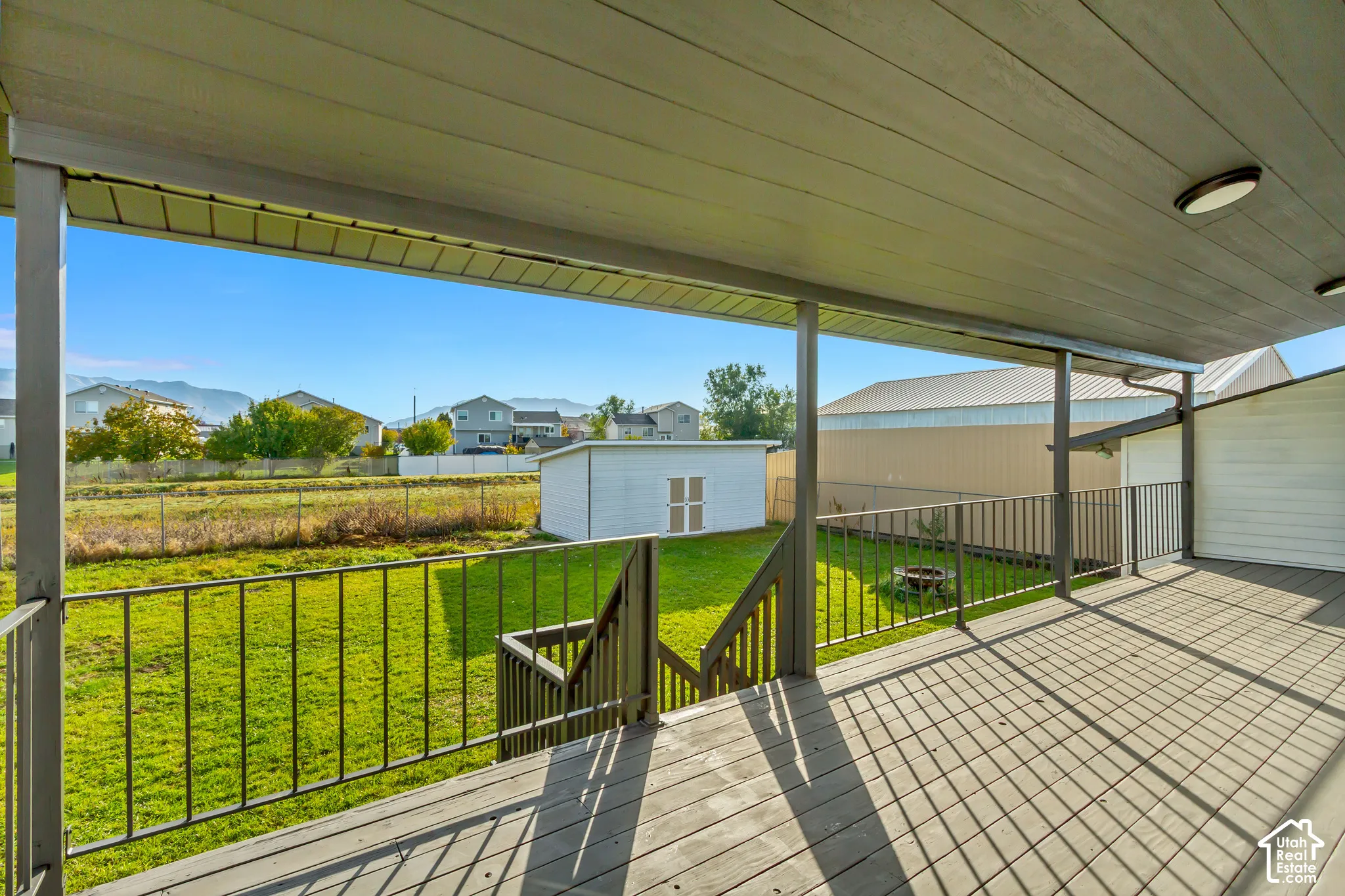 Covered exterior deck with metal railing and stairs, featuring an expansive backyard, detached storage shed, and mountain backdrop