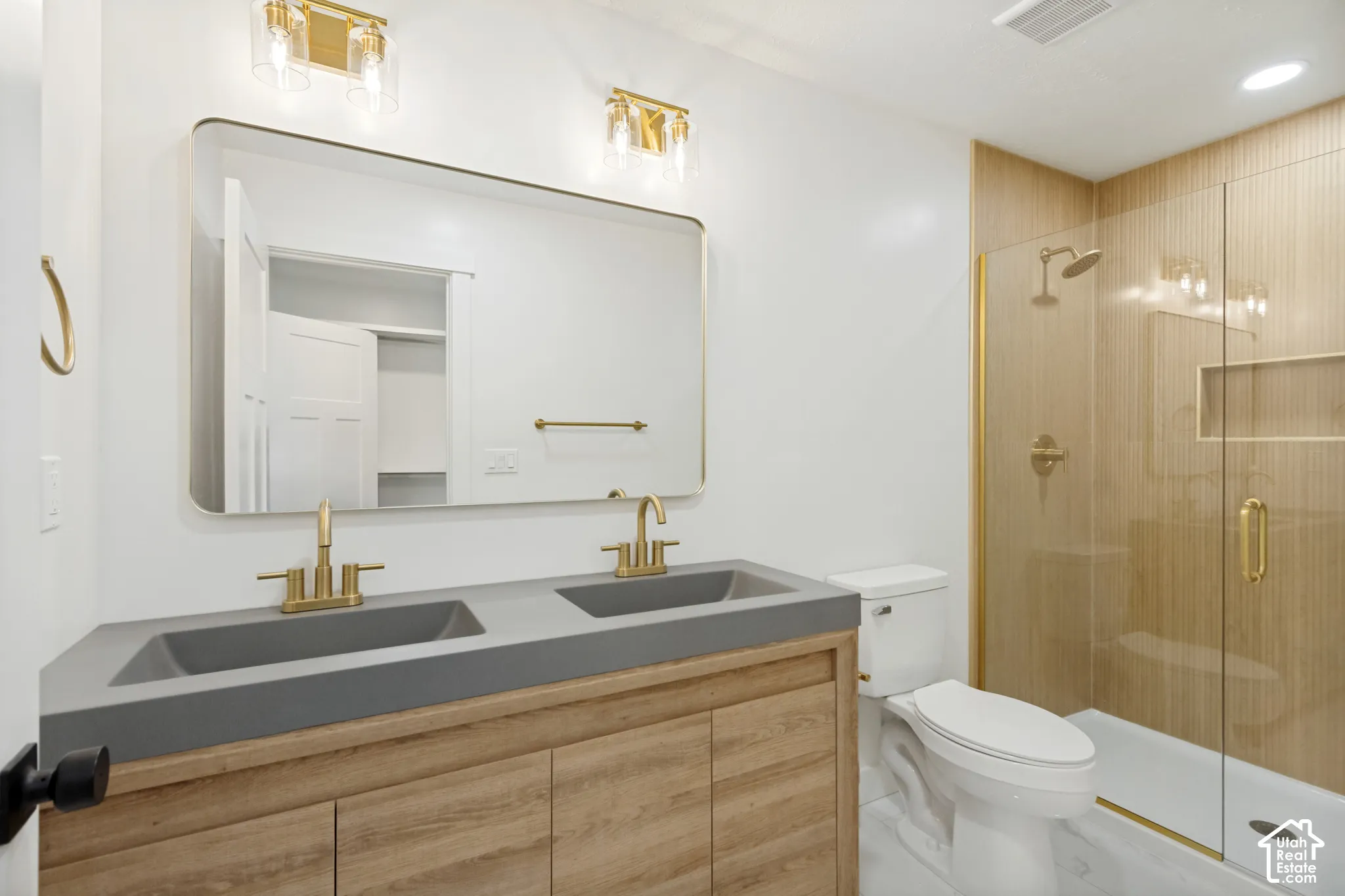 Double vanity with a matte-finish countertop and integrated sinks, brass-toned fixtures, wood-finish cabinetry, frameless glass shower enclosure, and a tiled shower surround with a built-in niche