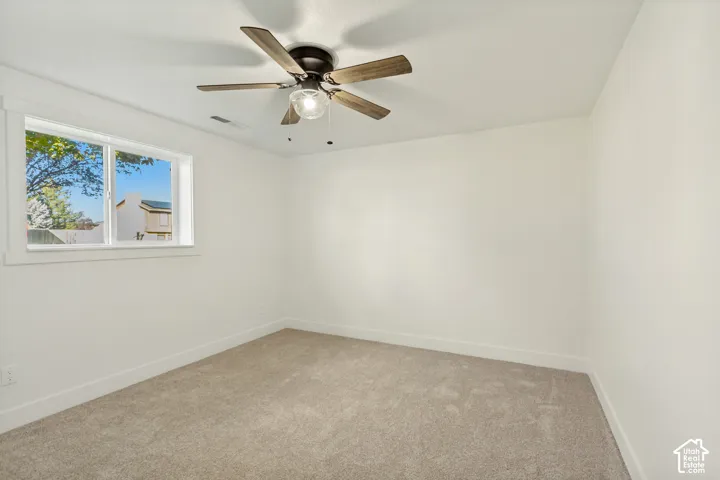 Neutral interior featuring light-toned wall-to-wall carpeting, a multi-blade ceiling fan with integrated light fixture, and a sliding window with wide white casing