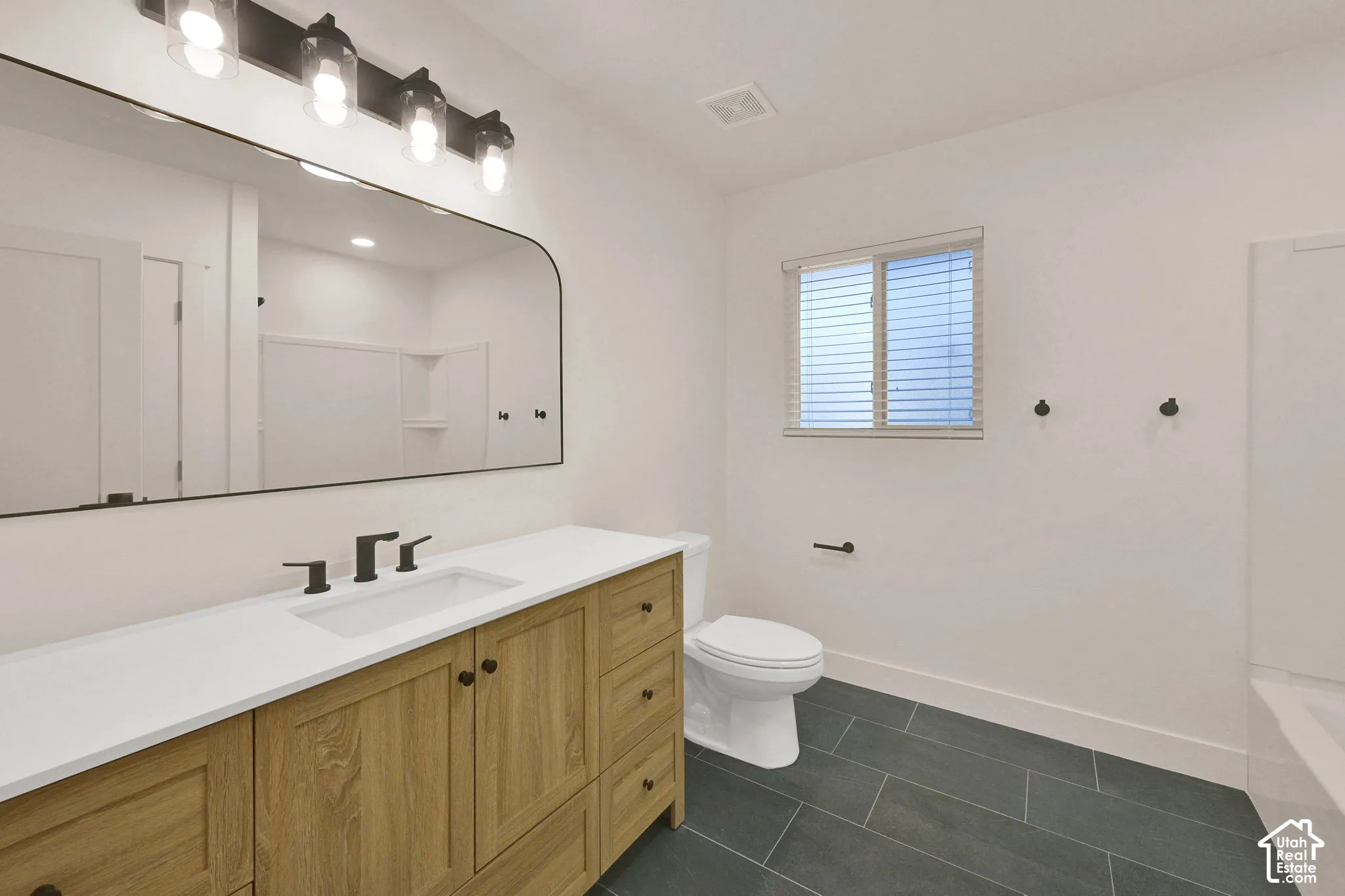 Wood-finish cabinetry featuring black hardware, white countertop with integrated rectangular sink, matte black faucet, large arched vanity mirror, and dark tile flooring
