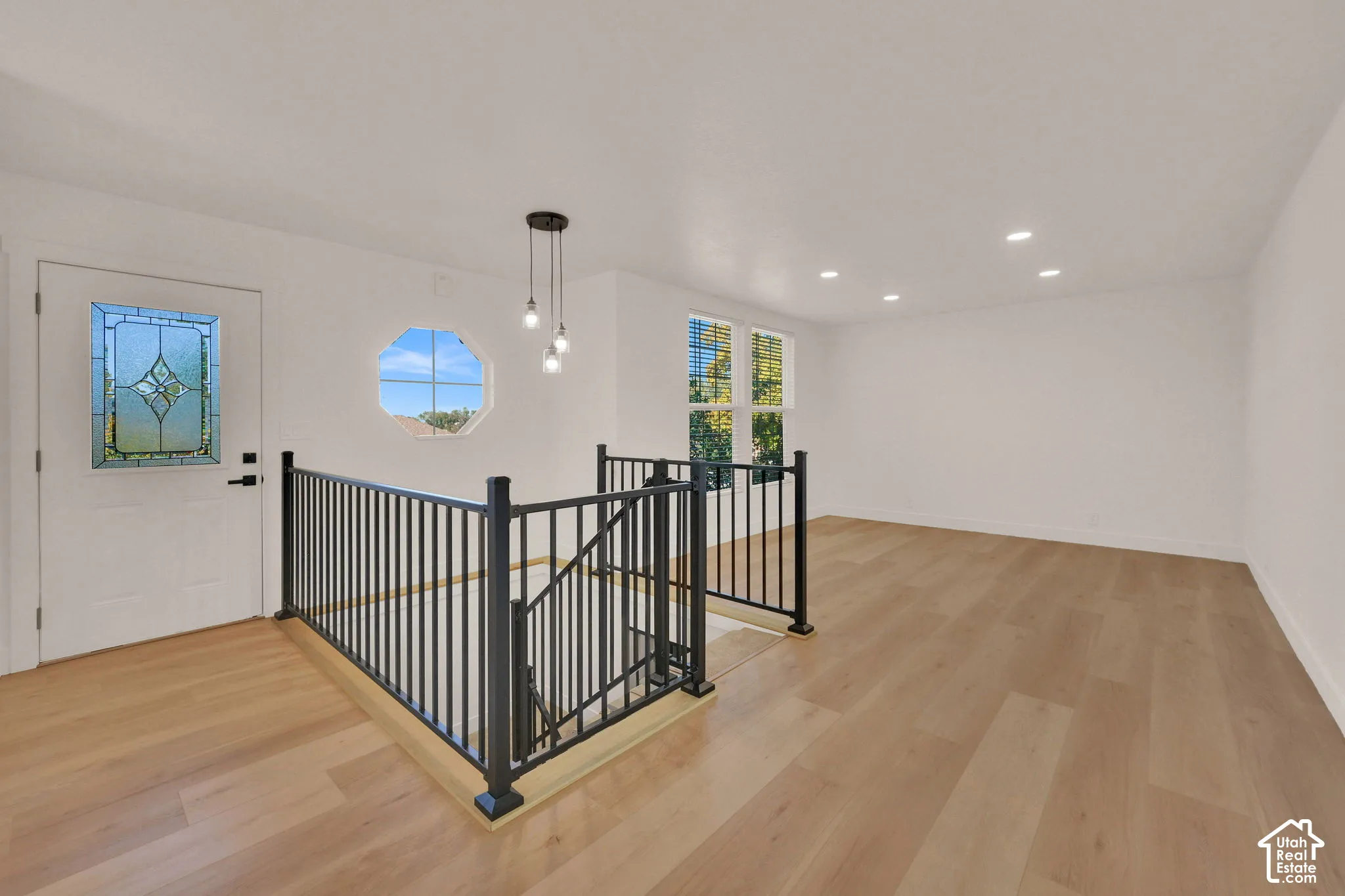 Open-concept interior featuring light wood-finish flooring, recessed lighting, matte black metal railing, octagonal window, and an entry door with decorative glass inlay