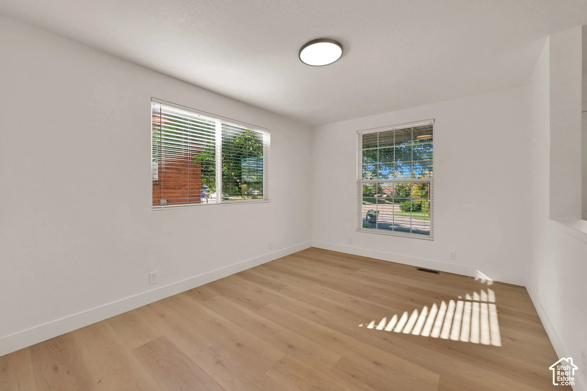 Interior space featuring wood-finish flooring, dual windows with fitted blinds, and a neutral white paint palette complemented by a flush-mount circular LED light fixture