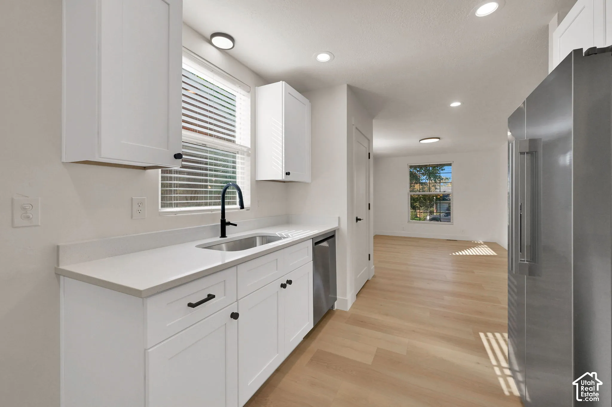 Kitchen featuring white shaker-style cabinetry, solid-surface countertops, a stainless steel refrigerator, matte black hardware, and light-toned wood-finish flooring