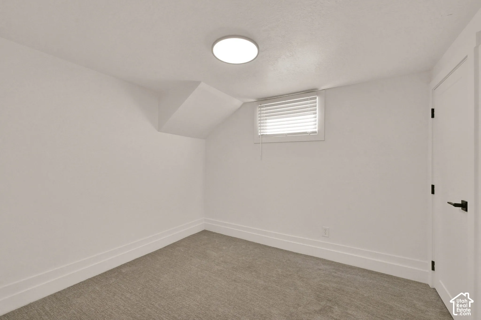 Interior space featuring neutral-toned carpeting, recessed ceiling lighting, a single horizontal window with fitted blinds, and a paneled door with matte black hardware