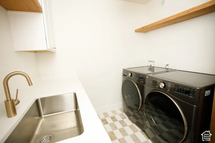 Laundry space featuring solid-surface countertops, an undermount stainless steel sink with a brass-finish faucet, white upper cabinetry, and natural wood floating shelving