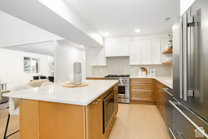 Kitchen featuring light-finish cabinetry, a central island with a solid-surface countertop, stainless steel appliances, a tile backsplash, and wood-finish flooring