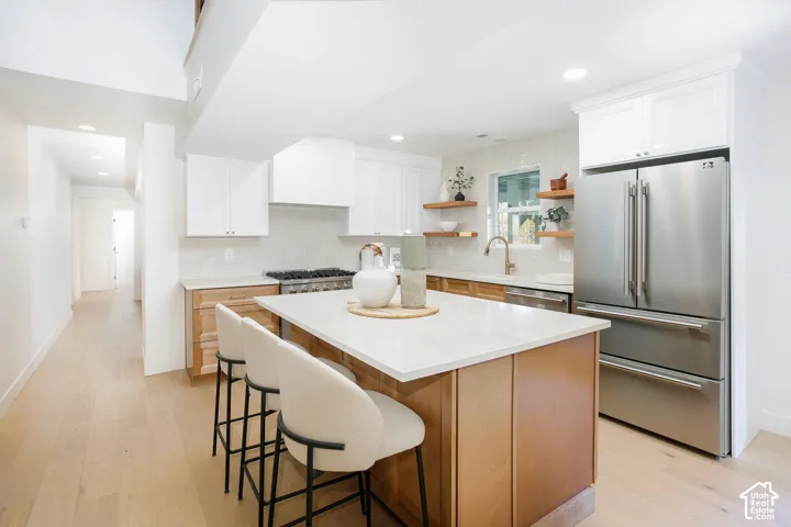 Kitchen featuring a central island with a solid surface countertop, white upper cabinetry, natural wood-finish lower cabinetry, stainless steel appliances, and floating wood shelves