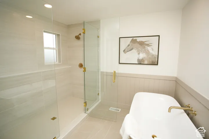 Walk-in shower with floor-to-ceiling tile, frameless glass enclosure, freestanding soaking tub, brass-finish fixtures, and wainscoting detail