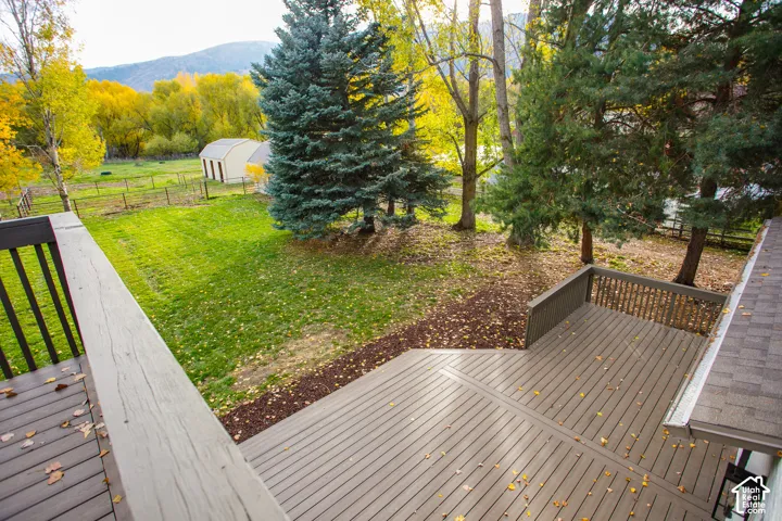 Multi-level wood-tone deck featuring vertical slat railings, spanning over a spacious yard with mature evergreen and deciduous trees, fenced perimeter, and a detached outbuilding against a mountain backdrop