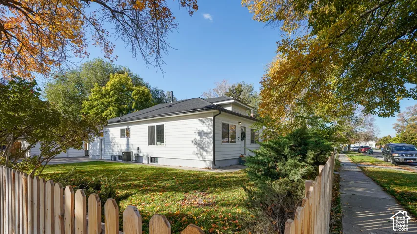 Single-story residence featuring white horizontal siding, a dark shingled roof, a front yard with mature trees, and a wood picket fence