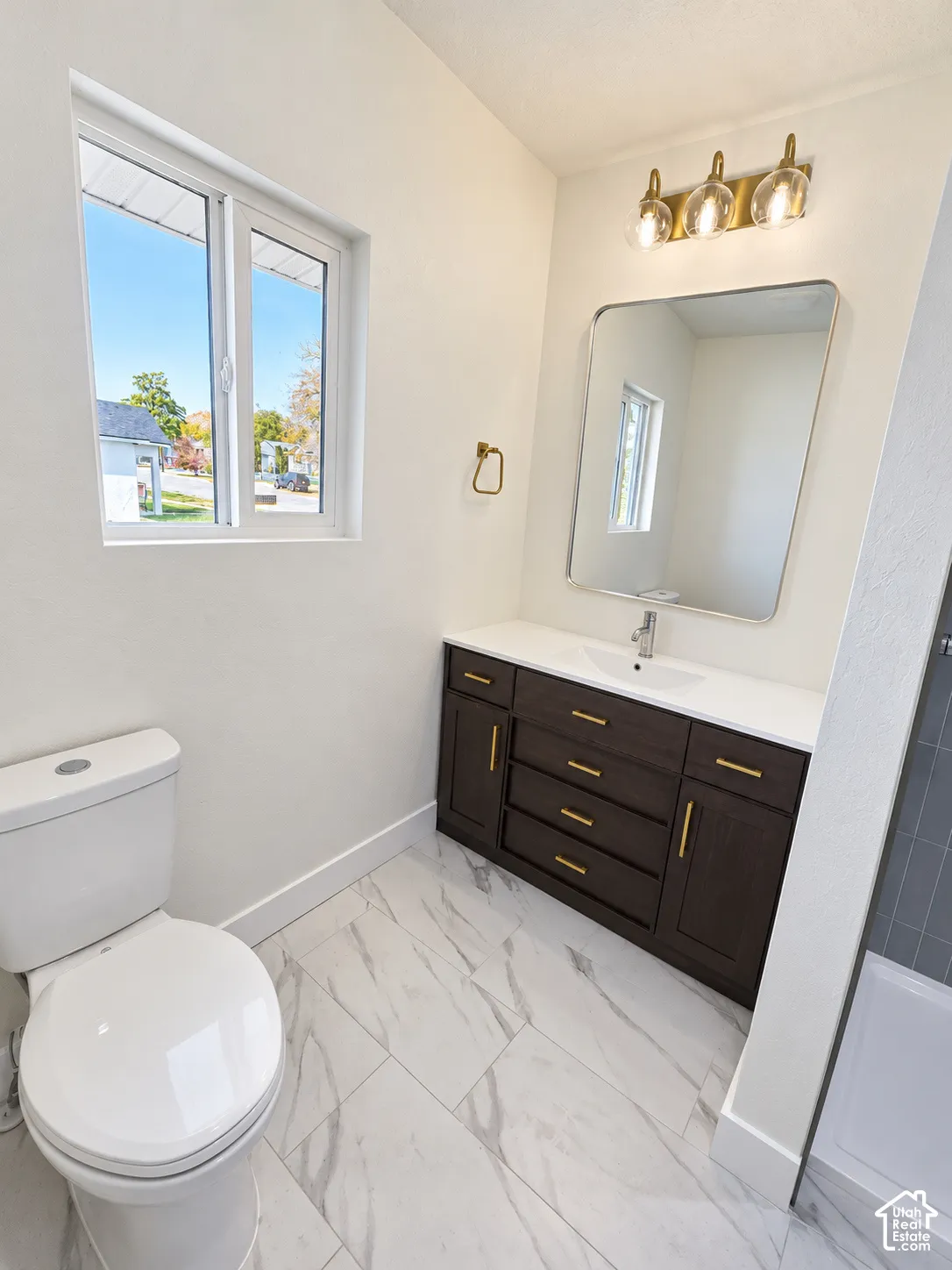 Bathroom featuring a dark wood-finish vanity with gold-tone hardware, white stone-composite countertop with an integrated basin, large-format tile flooring with marble-finish veining, and a three-light brass vanity fixture