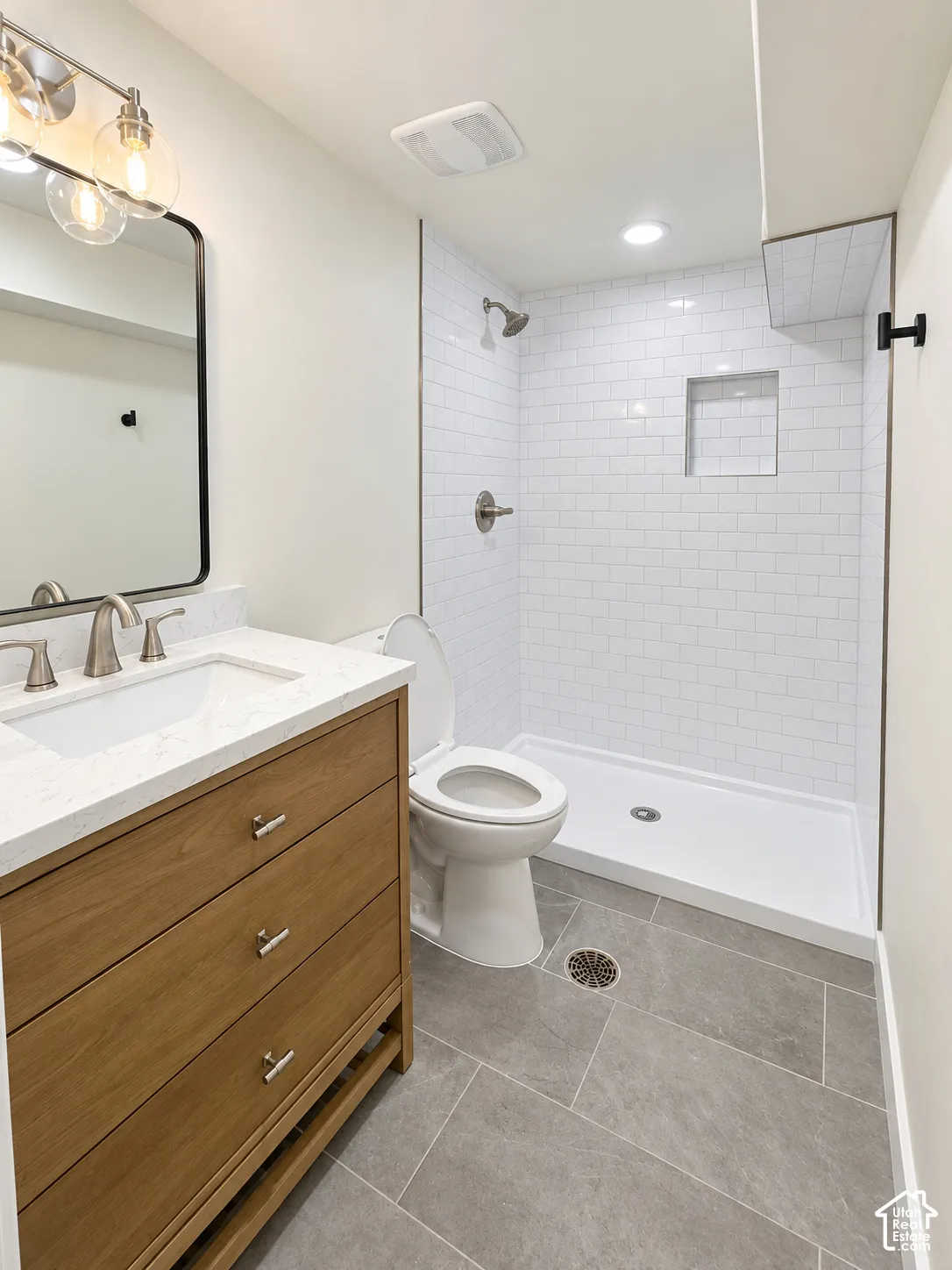 Bathroom featuring a wood-finish vanity with a stone-composite countertop, subway-tile shower surround with a built-in niche, and large-format tile flooring