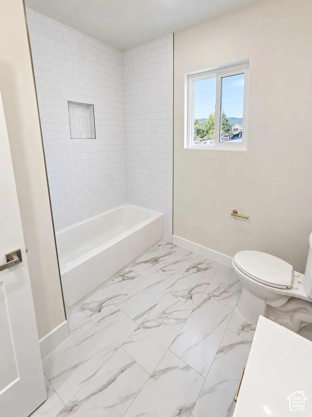 Bathroom featuring large-format tile flooring with a veined pattern, white subway tile tub surround with a recessed niche, and gold-finish hardware