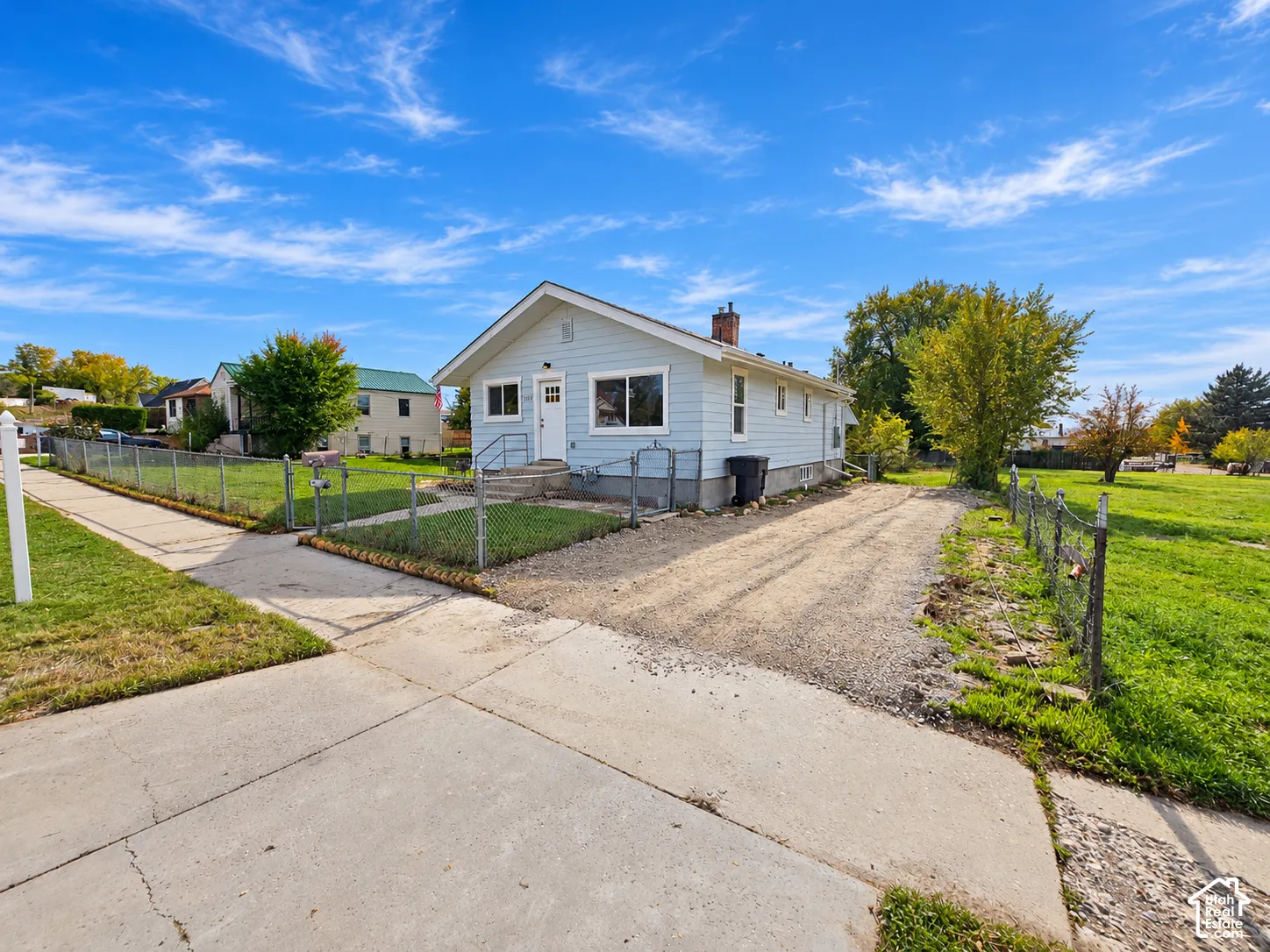 Single-story residence featuring light blue siding, a peaked roof with a masonry chimney, a gravel driveway, and a chain-link fenced perimeter