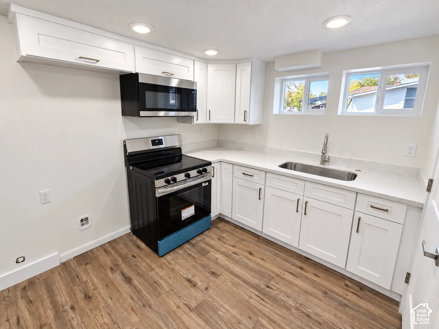 Kitchen featuring white shaker-style cabinetry, solid-surface countertops, wood-finish flooring, a stainless steel undermount sink, and black appliances including an over-the-range microwave and electric range