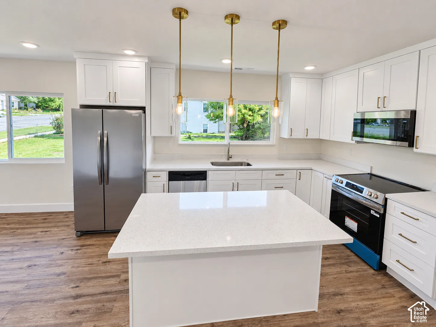 Kitchen featuring a central island with a solid surface countertop, shaker-style cabinetry, stainless steel appliances, and gold-tone pendant lighting over a wood-finish floor