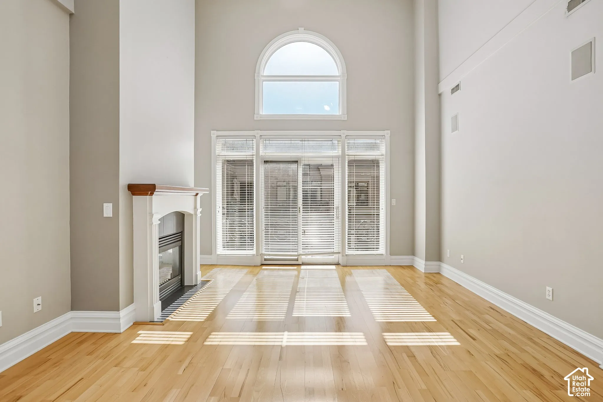 High-ceiling living area featuring wood-finish flooring, a prominent fireplace with a traditional mantle, and a large arched window above a glass sliding door