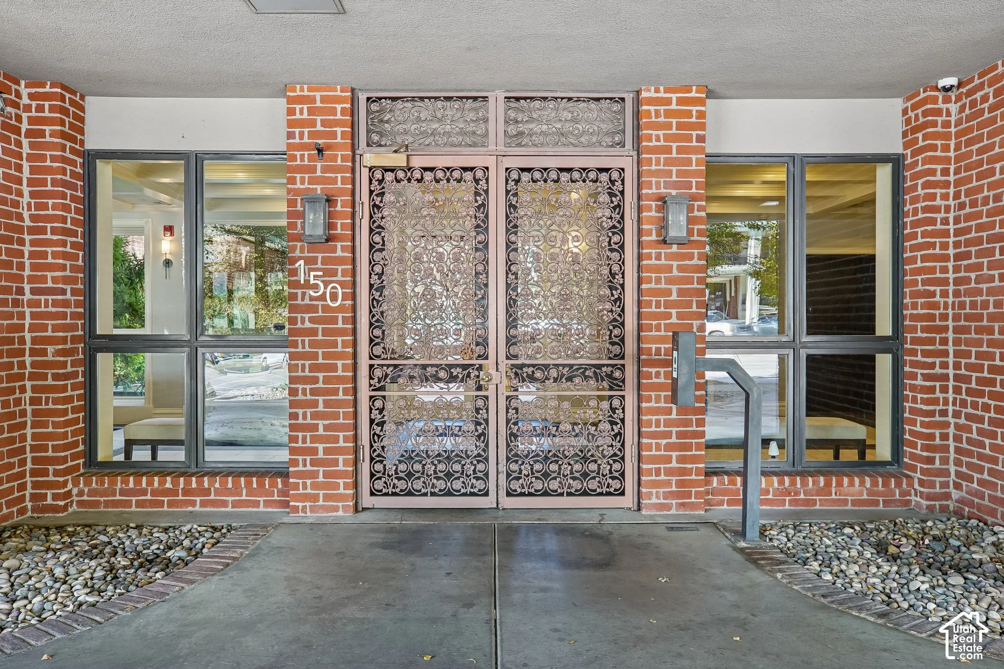 Exterior entrance featuring a red brick facade, double metal-grille doors, fixed-pane windows, and stone-bed landscaping