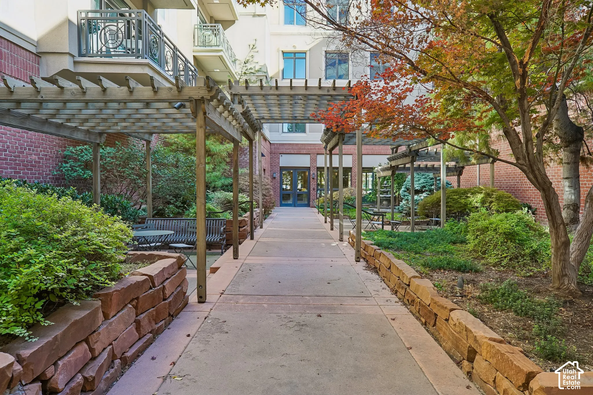 Courtyard walkway featuring timber pergolas, natural stone retaining walls, mature landscaping, and multi-story brick masonry exterior