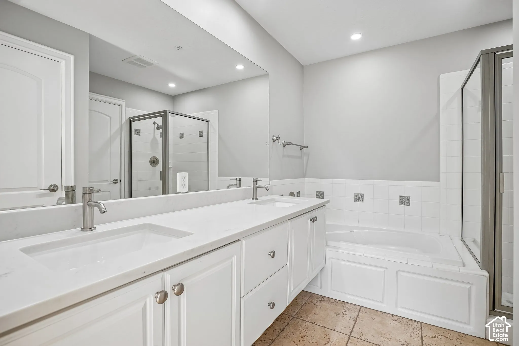 Dual-sink vanity with white cabinetry, light stone-finish countertops, recessed lighting, drop-in soaking tub, and separate glass-enclosed shower