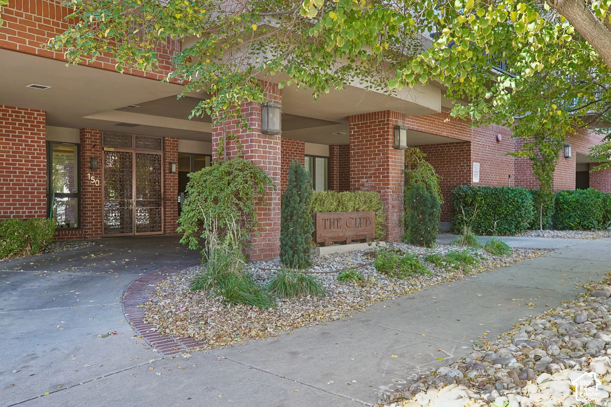 Building entrance featuring a brick facade, covered portico, decorative metal entry doors, and professional landscaping
