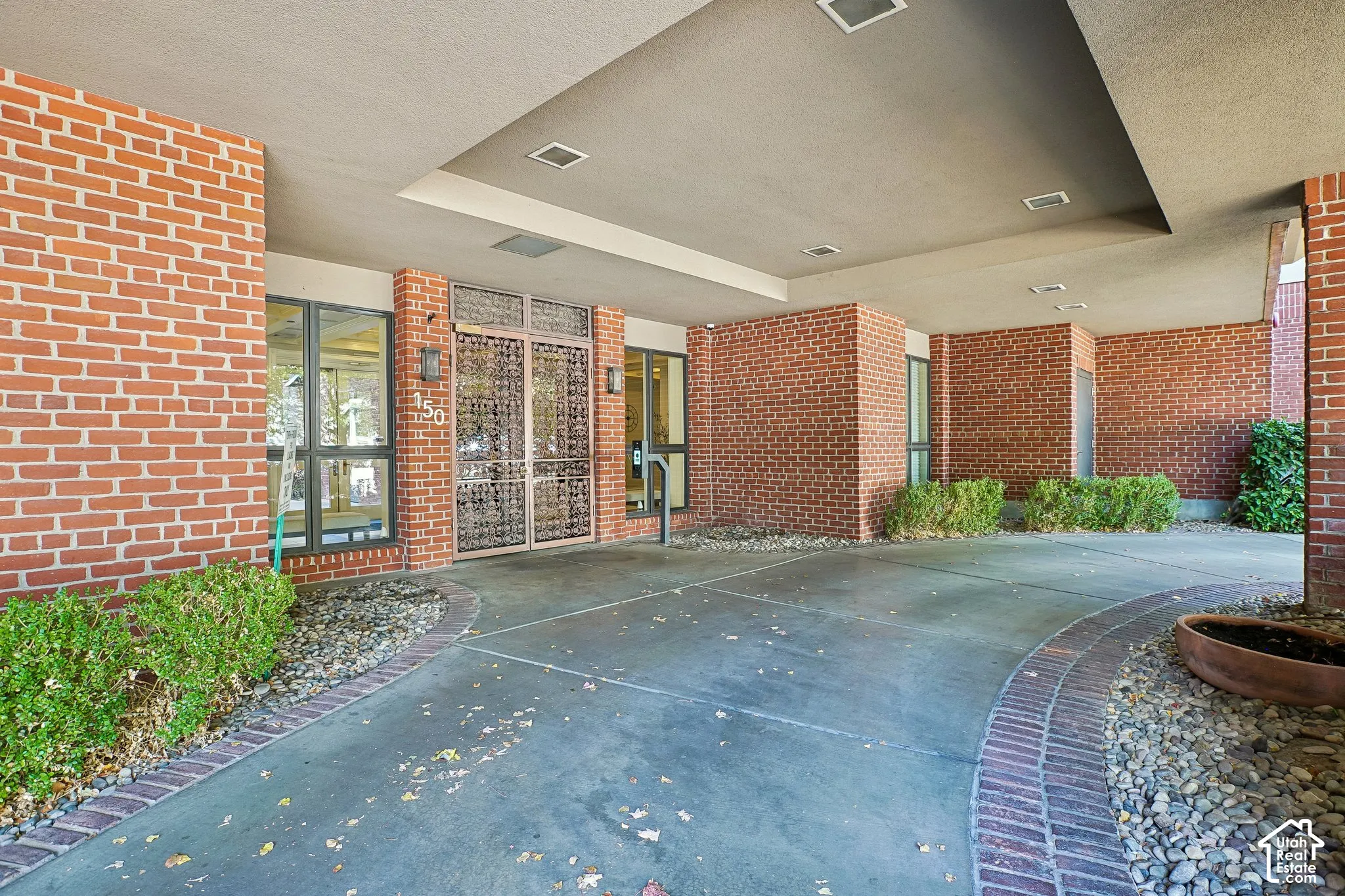 Covered entryway featuring a brick facade, recessed lighting, ornate metal double doors, and a concrete porte-cochere with brick border detailing