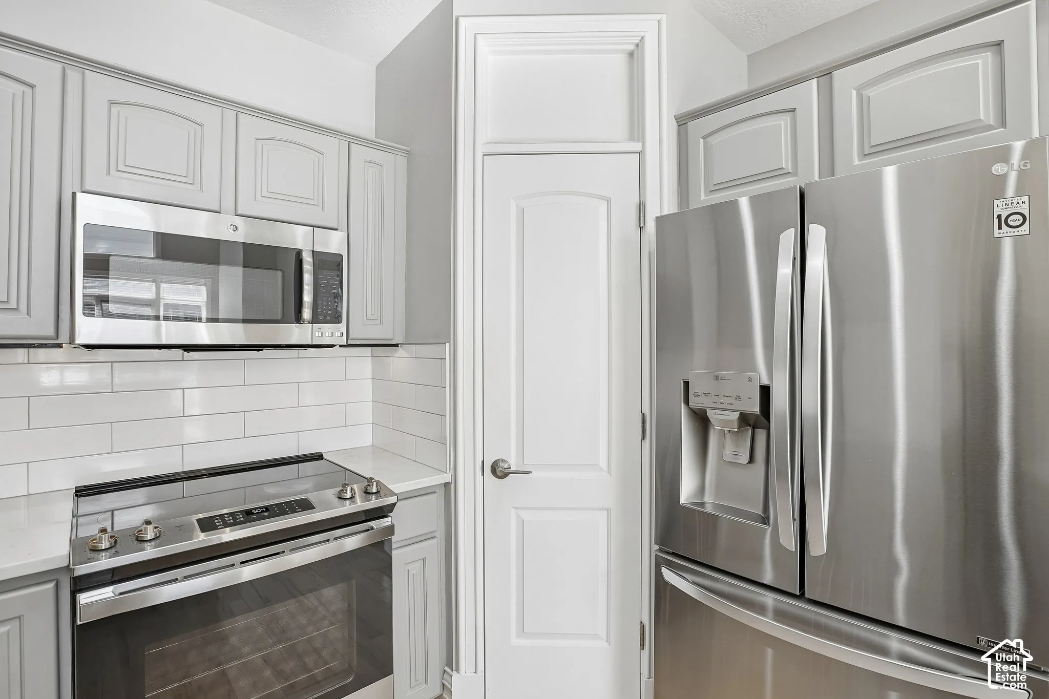 Kitchen featuring gray shaker-style cabinetry, stainless steel appliances, white subway tile backsplash, quartz countertops, and a full-height pantry door with crown molding