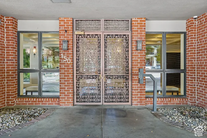 Exterior entrance featuring a red brick facade, double metal-grille doors, fixed-pane windows, and stone-bed landscaping