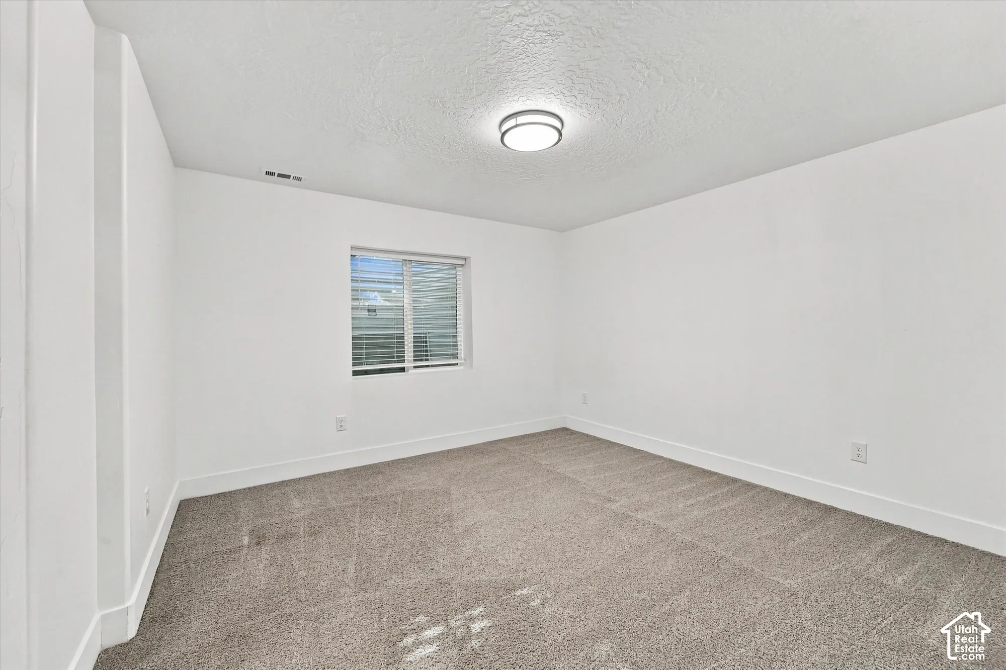 Neutral interior room featuring neutral-toned carpeting, white walls, a sliding window with integrated blinds, and a circular flush-mount ceiling light fixture