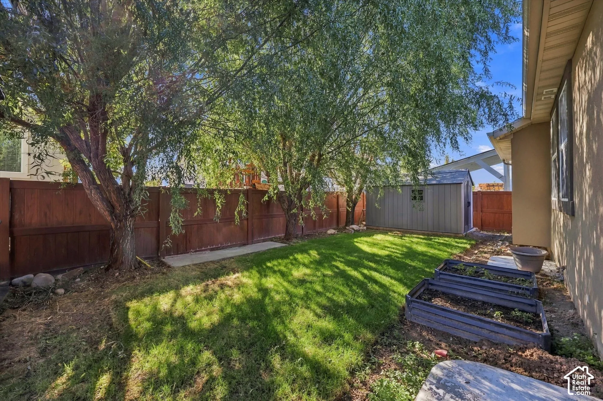 Backyard featuring a grass lawn, mature trees, wood-style perimeter fencing, a detached storage shed, and raised garden beds