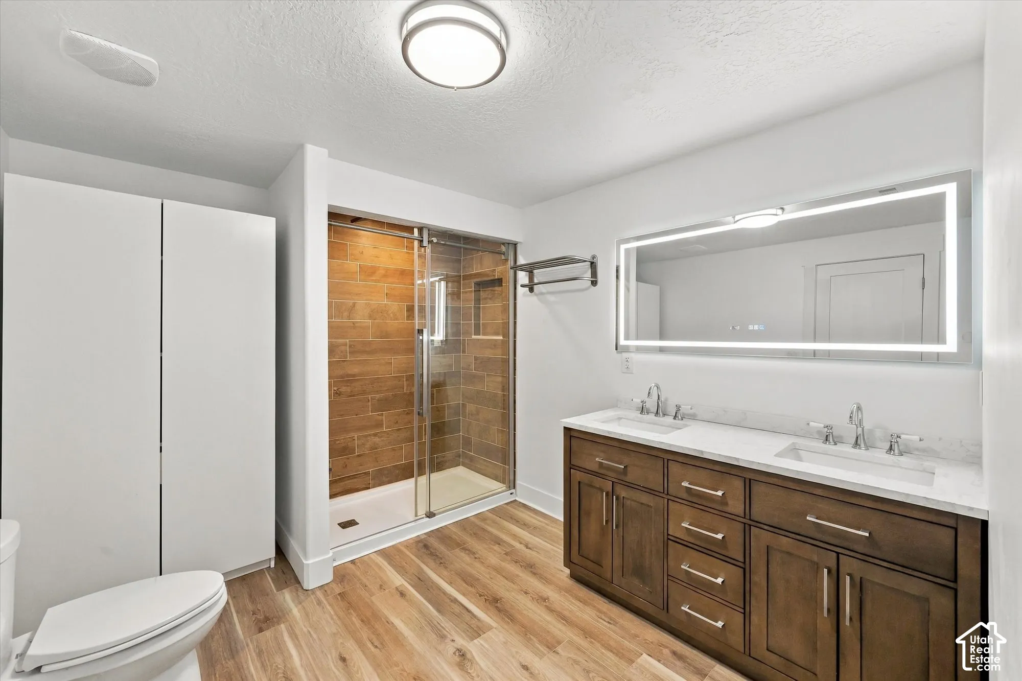Double vanity featuring a light-toned countertop, wood-finish cabinetry, a large integrated LED-lighted mirror, a glass-enclosed shower with wood-finish tile accents, and wood-finish flooring