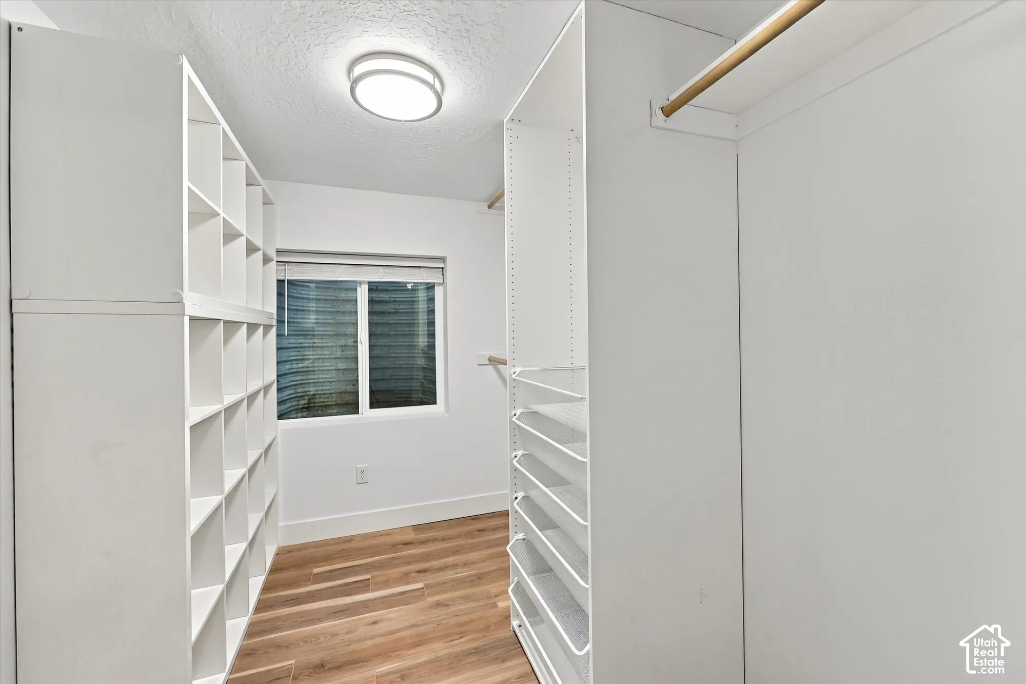 Walk-in closet featuring white floor-to-ceiling shelving units, tiered storage drawers, hanging rods, wood-finish flooring, and a flush-mount ceiling light