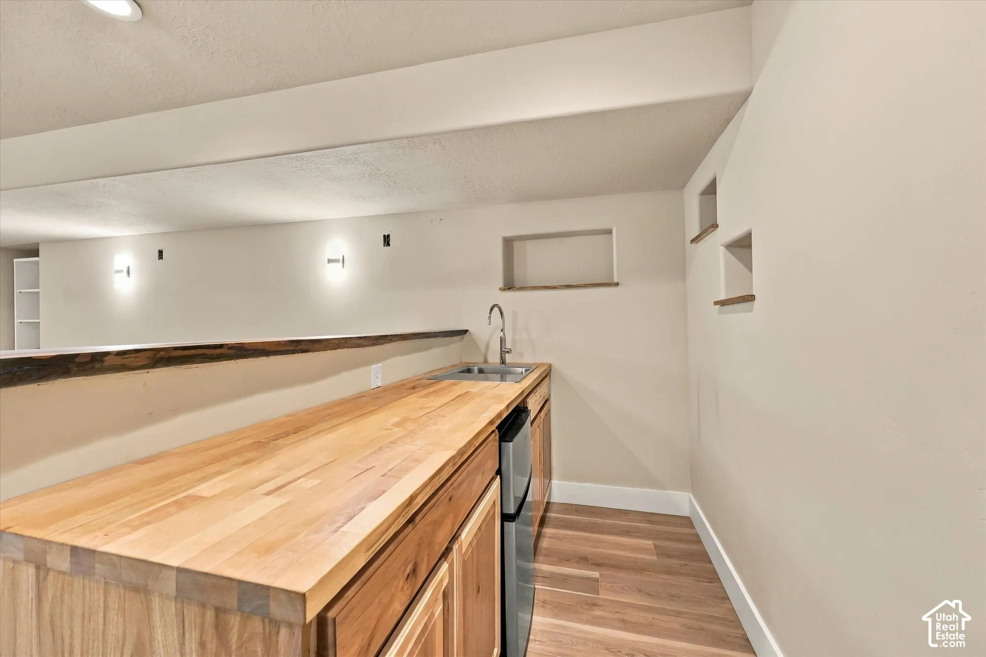 Wet bar featuring a butcher block countertop, wood cabinetry, stainless steel under-mount sink, integrated mini-refrigerator, and wall-mounted light fixtures