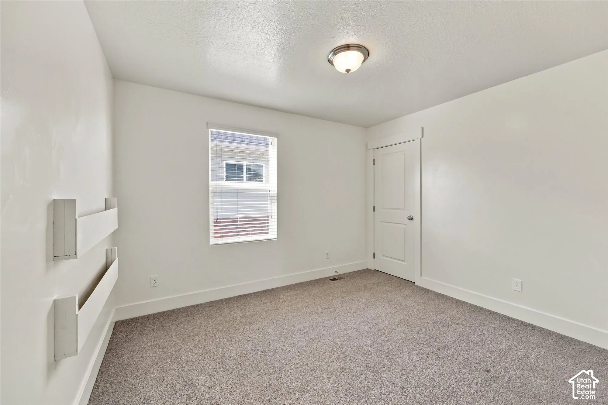 Interior room featuring neutral wall-to-wall carpeting, white walls, a single window with blinds, a recessed ceiling light fixture, and a built-in wall-mounted shelving unit